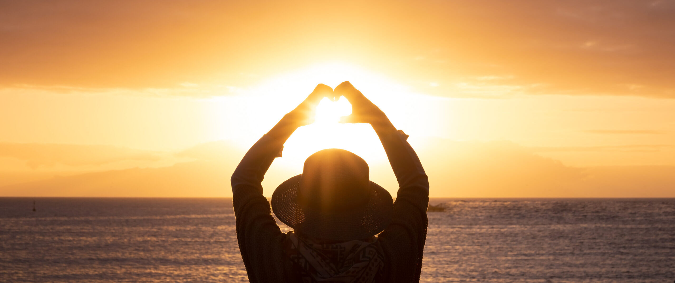 Rear view of woman silhouette admiring the orange sunset over the sea doing heart shape with her hands. Love and peace concept in beautiful nature